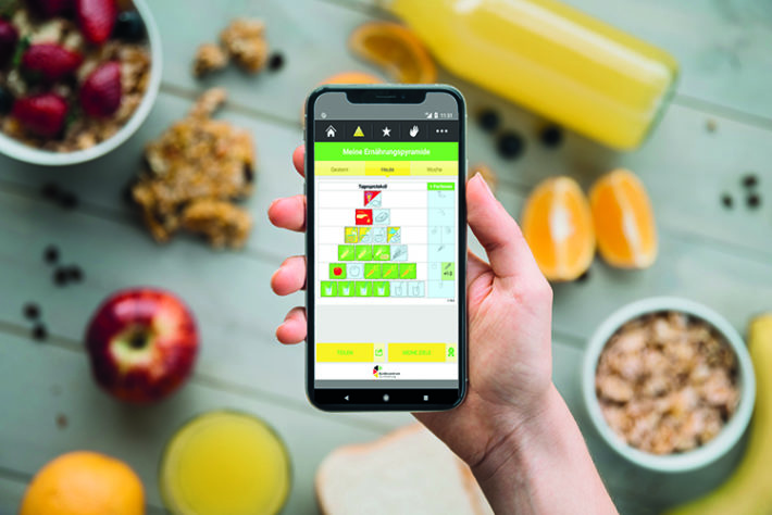 Eine Hand, die ein Handy hält auf dem die Ernährungspyramide abgebildet wird. Im Hintergrund liegen verschiedene Lebensmittel.