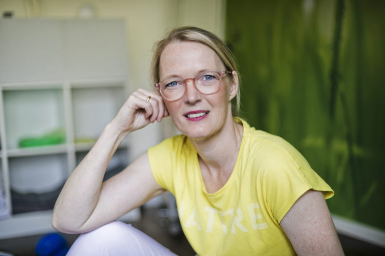 Porträt von Annette Rottempker, während sie ihren angewinkelten Arm auf ihrem Knie ablegt und ihren Kopf mit ihrer Hand stützt, vor unscharfem Hintergrund ihrer Praxis