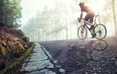 Rennradfahrer der durch an einer Waldstraße entlang fährt und einen Helm trägt. Die Sonne scheint