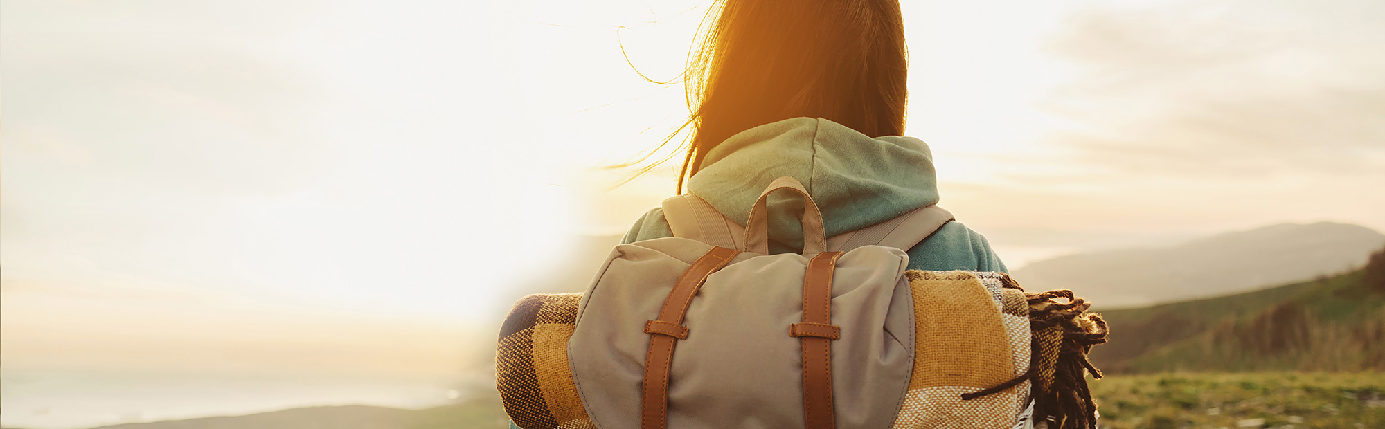 Zu sehen ist der Oberkörper einer Frau von hinten. Sie hat einen Rucksack auf und blickt in die Ferne. Die Sonne scheint ihr entgegen.