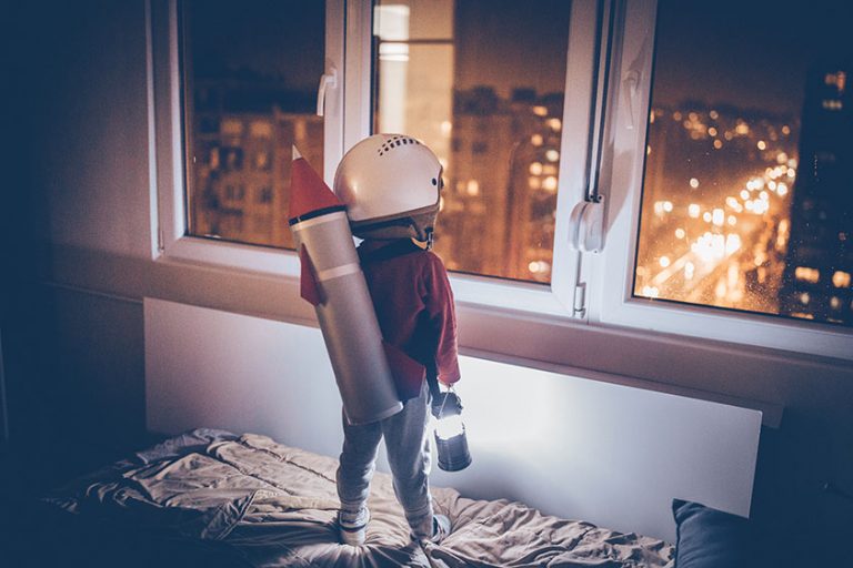 Bild eines kleinen Kindes von hinten, das einen Helm auf dem Kopf und eine kleine Rakete auf dem Rücken trägt, stehend auf dem Bett, aus dem Fenster schauend, mit einer kleinen Lampe in der Hand