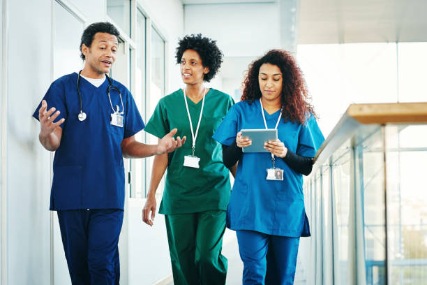Zu sehen sind drei Personen im Krankenhaus, die zusammen einen Gang lang gehen. Alle tragen unterschiedliche Kassacks. Ganz links läuft ein Mann in einem dunklen Kasack mit einem Stetoskop um den Hals. er erklärt irgendwas und macht mit seinen Händen Gestiken. in der Mitte läuft eine Frau in einem dunkel grünen Kasack, sie hört dem Arzt zu. ganz links läuft eine Frau in einem hell blauen Kasack. Sie hat ein Tablett in der Hand.