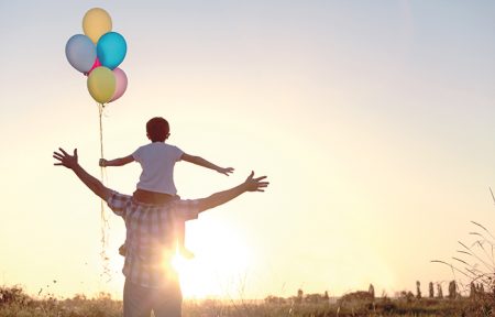 Zu sehen ist ein Mann der ein Kind auf den Schultern trägt. Beide breiten ihre Arme zur Seite aus und gucken in die Sonne. Das Kind hält mehrere bunte Luftballons in der Hand. Sie stehen auf einem Feld.